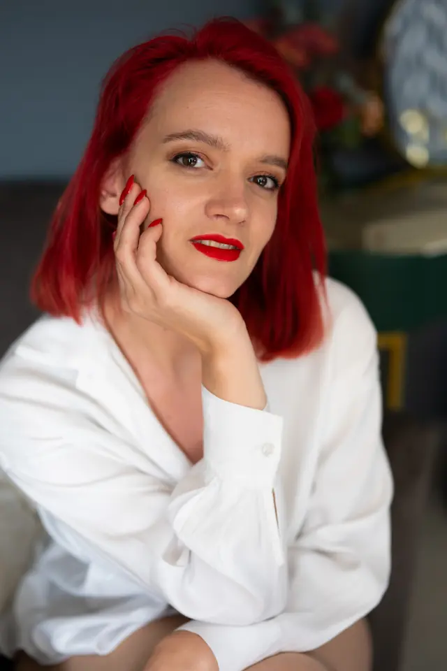 Vrouw met rood haar en rode lippen in een witte blouse, kin op haar hand